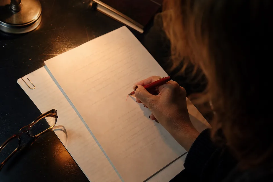A hand marking corrections on a printed page with a red pen in lamp light